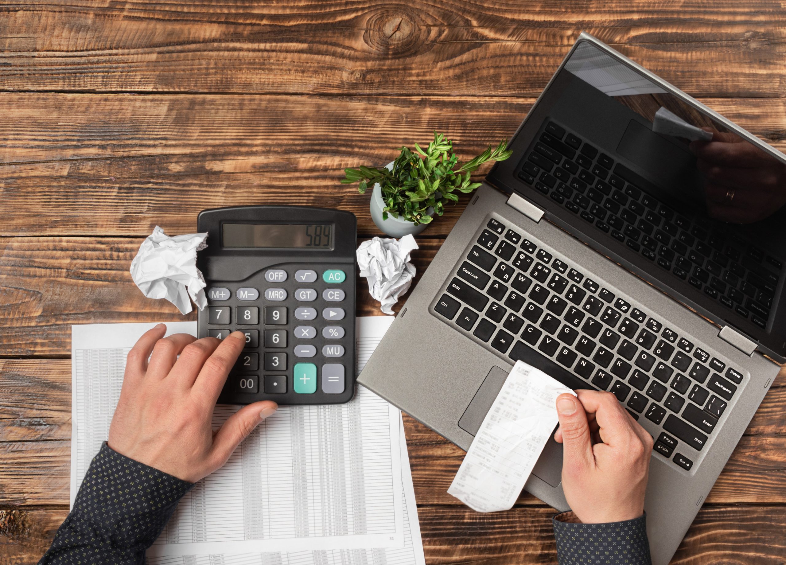 Male hand holding a check and using a calculator when filling out an individual tax return, close. Tax calculation, account, income and expense analysis, accounting. Wooden table flat lay copy space.